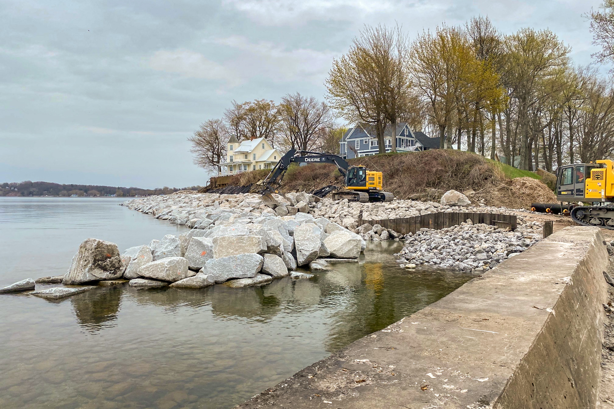 an excavator moving large seawall rock next to homes on a peninsula